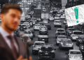 Colapso-vial-definitivo-de-nuevo-leon - Vecinos Unidos Blurry Man in a Suit in the Foreground; Congested City Street Full of Cars Behind; Torn White Paper with Teal Exclamation Mark at Top-right. - Vecinos Unidos