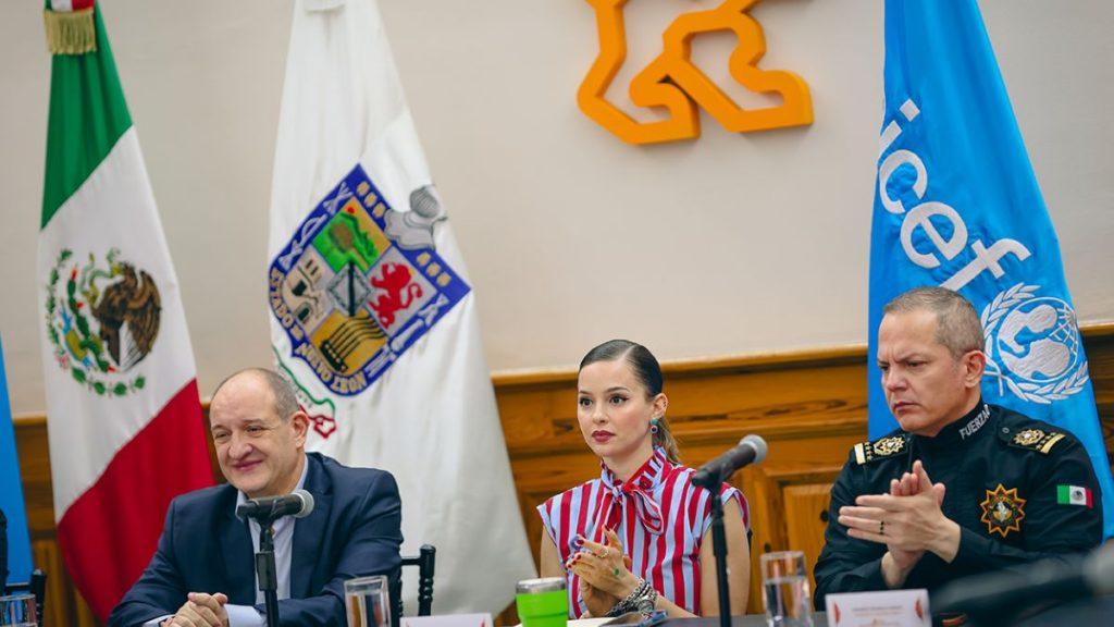 Mariana-rodriguez-uso-a-unicef-para-hacer-campana - Vecinos Unidos Panel Discussion with Three Speakers at a Table, Mexican and Un Flags in the Background and Microphones Before Them. - Vecinos Unidos
