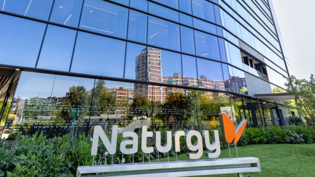 Exterior View of a Glass-front Office Building with a Large Naturgy Sign and Orange Leaf Logo, Reflecting Surrounding Buildings in the Windows, with Greenery in the Foreground. - Vecinos Unidos