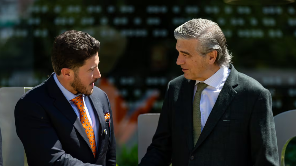 Two Men in Suits Shake Hands and Talk Outdoors, One in a Blue Suit with an Orange Polka-dot Tie and Pocket Square, the Other in a Dark Blazer with a Green Tie. - Vecinos Unidos