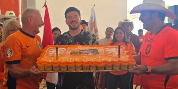 Group of People in Orange Sports Shirts Holding a Large Rectangular Orange Cake, Celebrating Together Indoors, with Flags in the Background and Cheering Faces Behind Them. - Vecinos Unidos