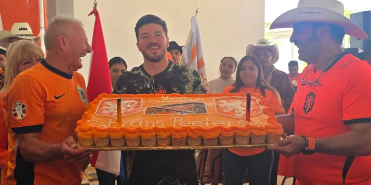 Group of People in Orange Sports Shirts Holding a Large Rectangular Orange Cake, Celebrating Together Indoors, with Flags in the Background and Cheering Faces Behind Them. - Vecinos Unidos