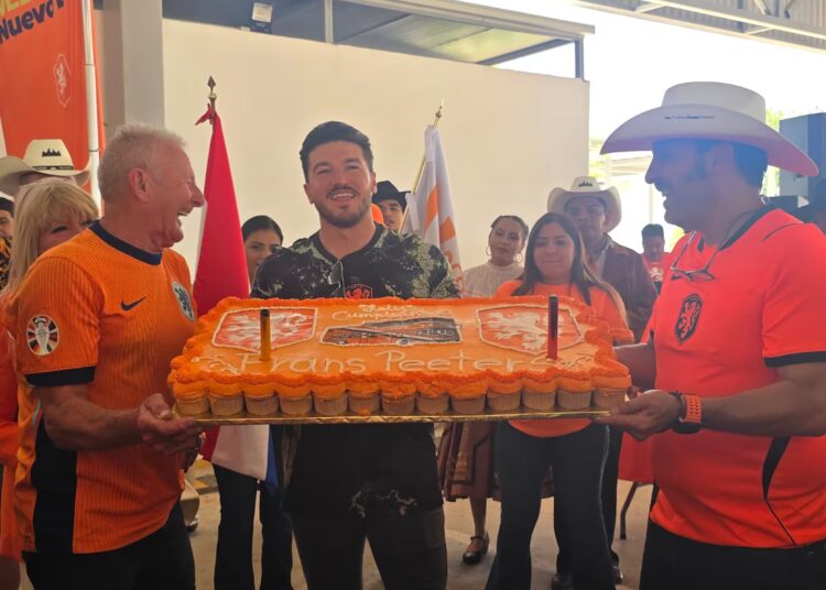 Group of People in Orange Sports Shirts Holding a Large Rectangular Orange Cake, Celebrating Together Indoors, with Flags in the Background and Cheering Faces Behind Them. - Vecinos Unidos