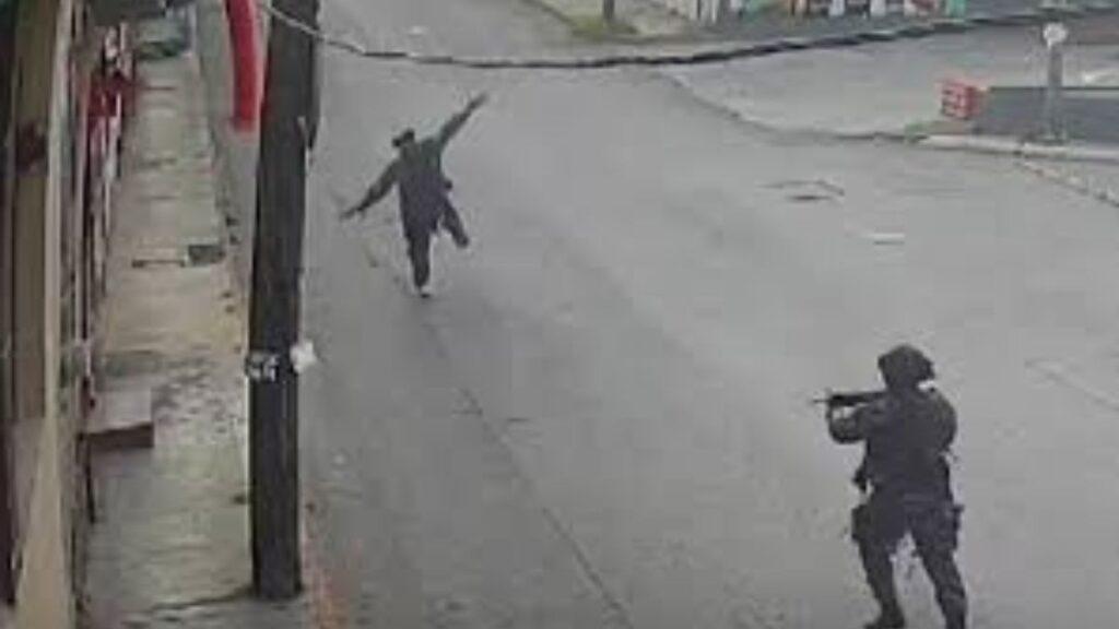 Two People on a Quiet City Street: a Person in Dark Clothing Runs with Arms Out, While Another Person in Tactical Gear Points a Rifle Toward Them on the Right Side of the Frame. - Vecinos Unidos