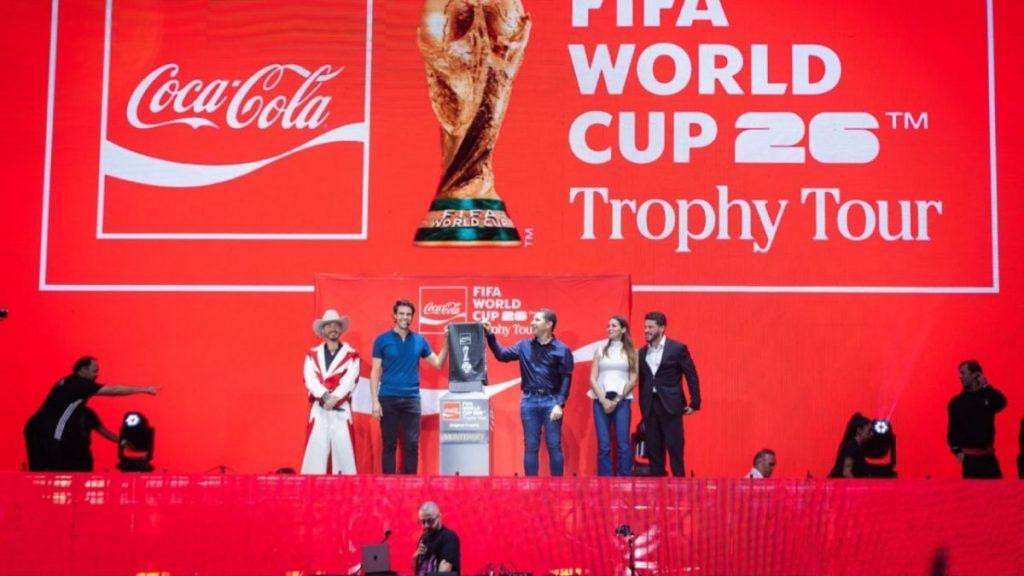 El-ambiente-en-el-fan-fest-de-nuevo-leon-tras-el-abucheo-al-gobernador - Vecinos Unidos Red Stage with Large Fifa World Cup Trophy Tour Branding and Coca-cola Logo; a Trophy on a Pedestal Center Stage with Several People Posing Nearby, Including a Man in Blue Shirt and a Woman in White Top. - Vecinos Unidos