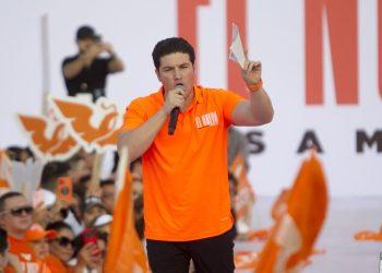 Man in Orange Polo Speaks into a Microphone at a Crowded Rally with Orange Flags and Banners Behind Him. - Vecinos Unidos