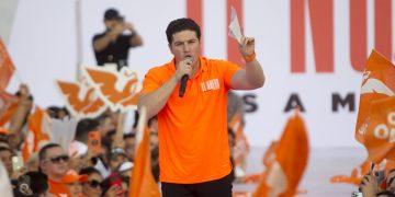 Man in Orange Polo Speaks into a Microphone at a Crowded Rally with Orange Flags and Banners Behind Him. - Vecinos Unidos